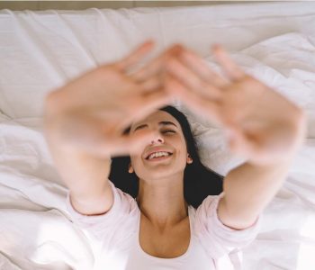 Dekoratives Bild einer Frau in einem Bett, welche die Augen geschlossen hat und lachend die Hände nach oben streckt.